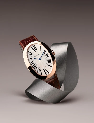 Elegant rose gold Cartier watch with a white face and brown leather strap, showcased against a silver ribbon and a soft grey background.