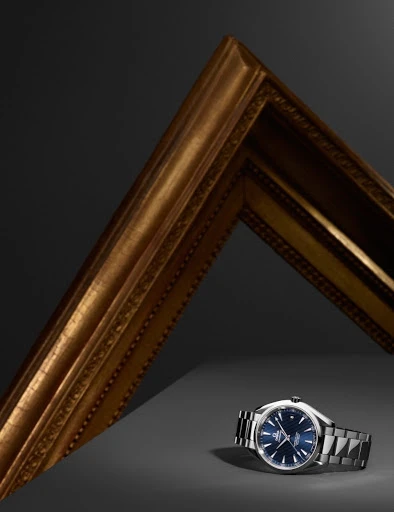 Wristwatch with a blue dial and a metallic strap sits beside a gold picture frame against a dark gray backdrop, spotlighted for effect.