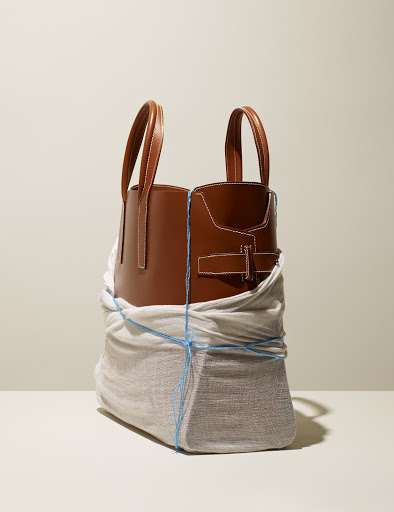 Studio shot of a brown leather tote bag partially wrapped in cloth and secured with blue string, set against a neutral background.