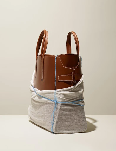 Studio shot of a brown leather tote bag partially wrapped in cloth and secured with blue string, set against a neutral background.