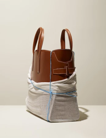 Studio shot of a brown leather tote bag partially wrapped in cloth and secured with blue string, set against a neutral background.