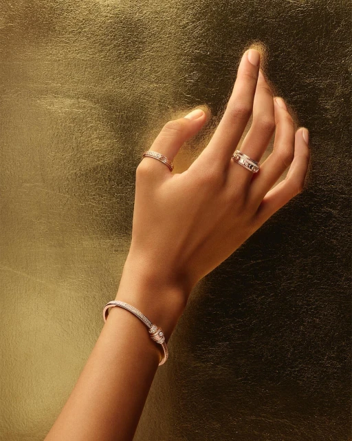 Close-up of a woman's hand with three rings and a bracelet against a metallic gold background, showcasing fine jewelry.