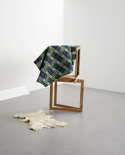 Still life of a geometric composition featuring a blue and green plaid textile draped over wooden frames, with a sheepskin on the floor.