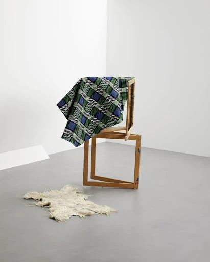 Still life of a geometric composition featuring a blue and green plaid textile draped over wooden frames, with a sheepskin on the floor.