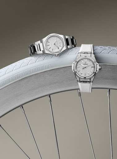 Two luxury silver wristwatches with diamond detailing are displayed on a white bicycle tire against a plain studio backdrop. 
