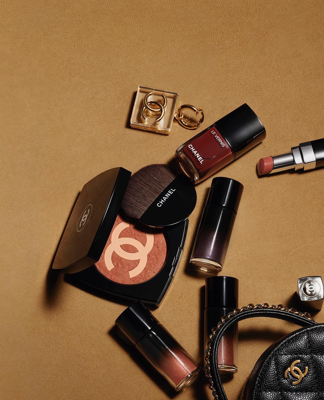 Chanel makeup still life: compact, blush, brush, lipstick, lip gloss, nail polish, rings, and purse on a tan surface.