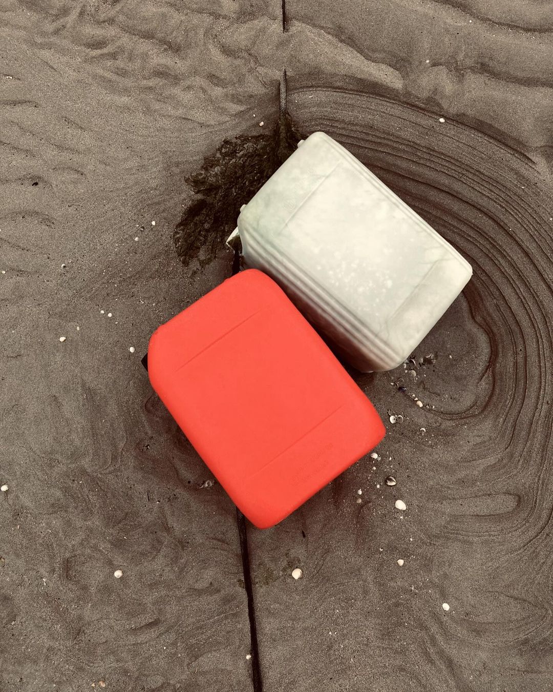 Two plastic containers sit on a dark sandy beach. One is bright red; the other is a light grey/green. The sand is a dark grey-brown color.