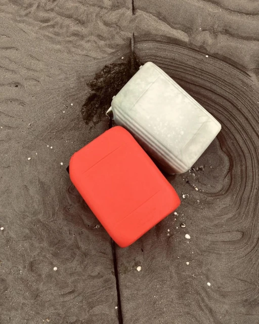 Two plastic containers sit on a dark sandy beach. One is bright red; the other is a light grey/green. The sand is a dark grey-brown color.
