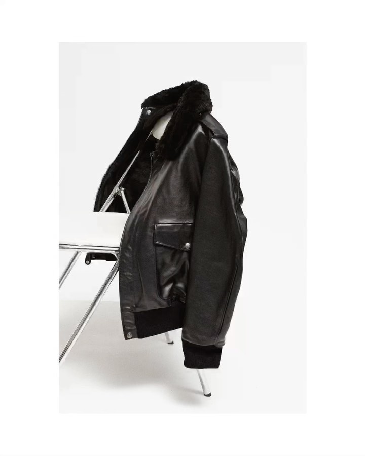Black leather bomber jacket with fur collar on a white wire chair in a bright, neutral studio setting. Fashion product shot.