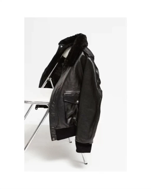 Black leather bomber jacket with fur collar on a white wire chair in a bright, neutral studio setting. Fashion product shot.