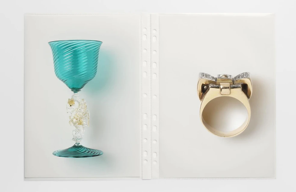 Elegant still life features a turquoise glass goblet with decorative stem and a gold ring with diamonds against a plain white backdrop.