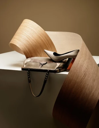 High-heeled shoe and handbag resting on a white surface with a curling wooden ribbon element against a warm neutral background.