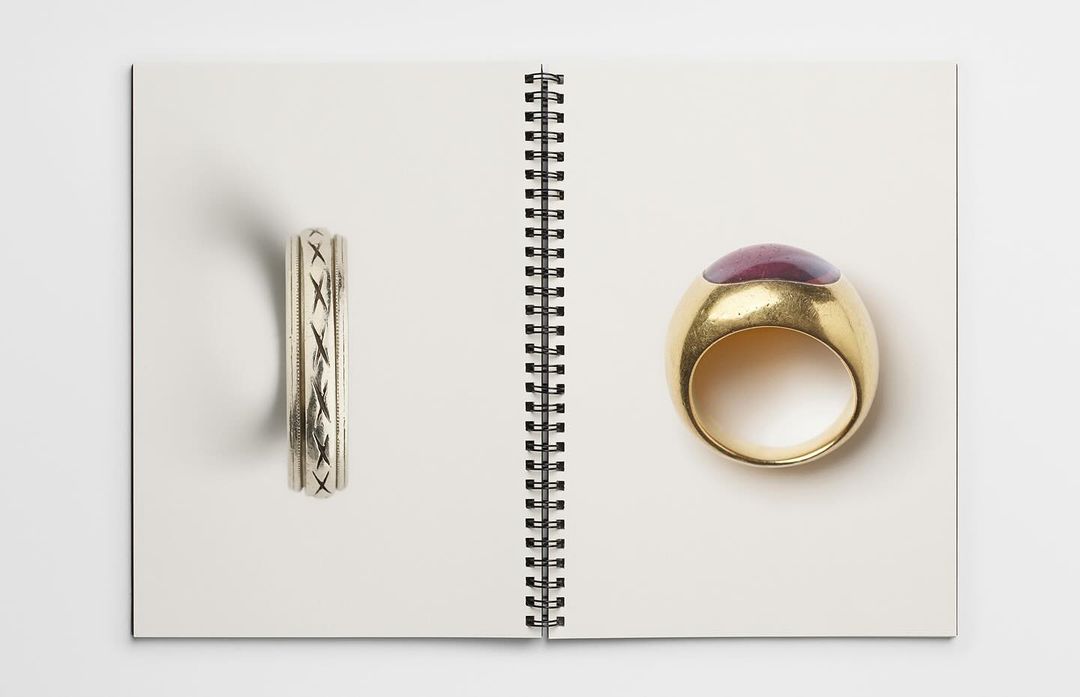 Two rings are displayed open on the pages of a white spiral-bound notebook, one silver with inlaid design, and another gold with a purple stone.