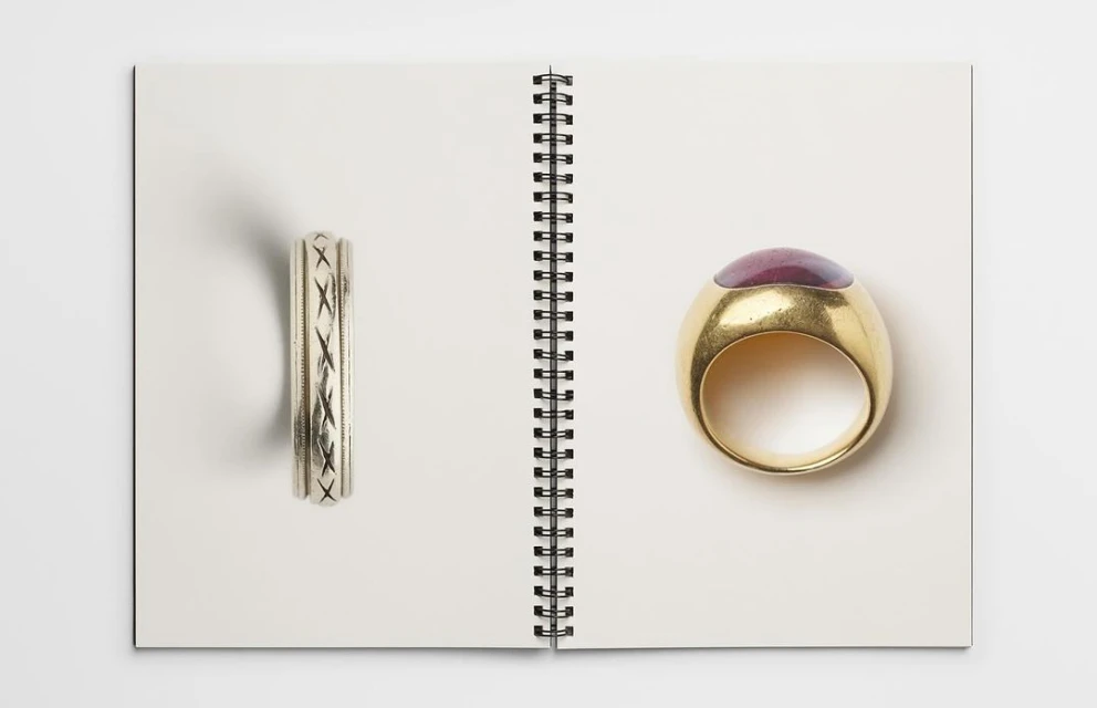 Two rings are displayed open on the pages of a white spiral-bound notebook, one silver with inlaid design, and another gold with a purple stone.