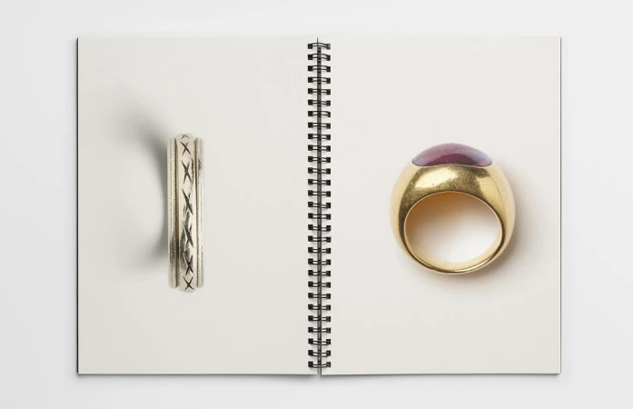 Two rings are displayed open on the pages of a white spiral-bound notebook, one silver with inlaid design, and another gold with a purple stone.