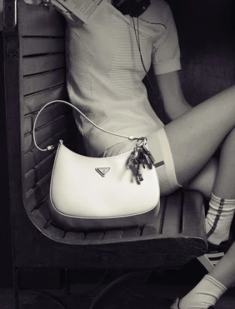 Monochrome fashion editorial image featuring a white shoulder bag resting on a wooden slatted bench next to a seated model.