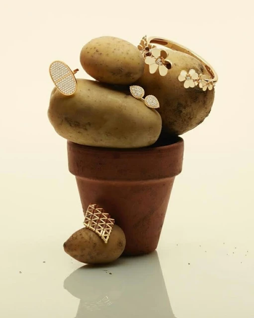 Still life of gold jewelry artfully arranged on and around a brown terracotta pot filled with a stack of fresh potatoes.