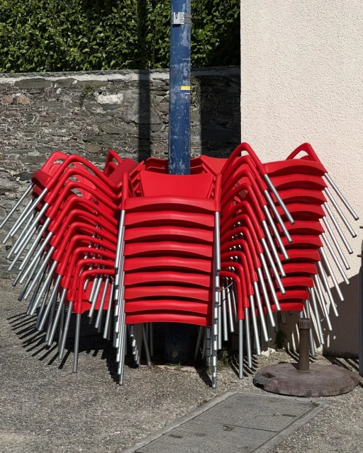 Stack of bright red chairs with silver legs piled up outdoors against a textured wall next to a blue metal pole.