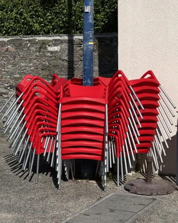 Stack of bright red chairs with silver legs piled up outdoors against a textured wall next to a blue metal pole.