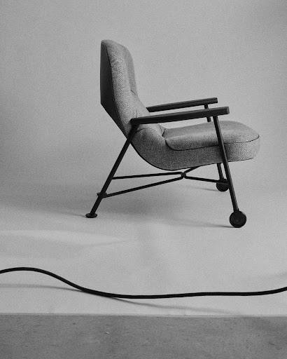 Monochrome image of a modern gray upholstered chair with dark wood armrests and a thin black metal frame with attached wheels.