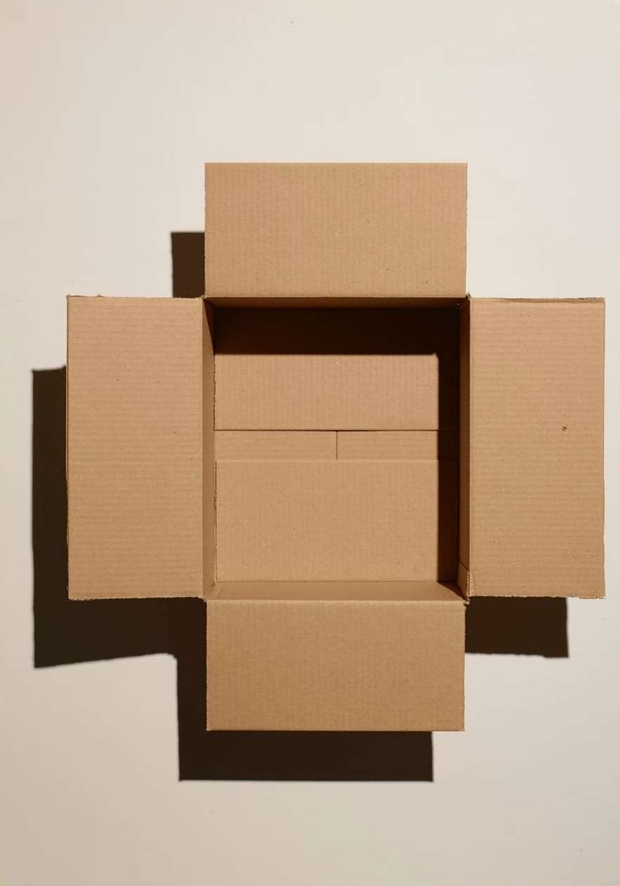 Open brown cardboard box with flaps extended, casting shadows on a neutral background, suitable for packaging or storage concepts.