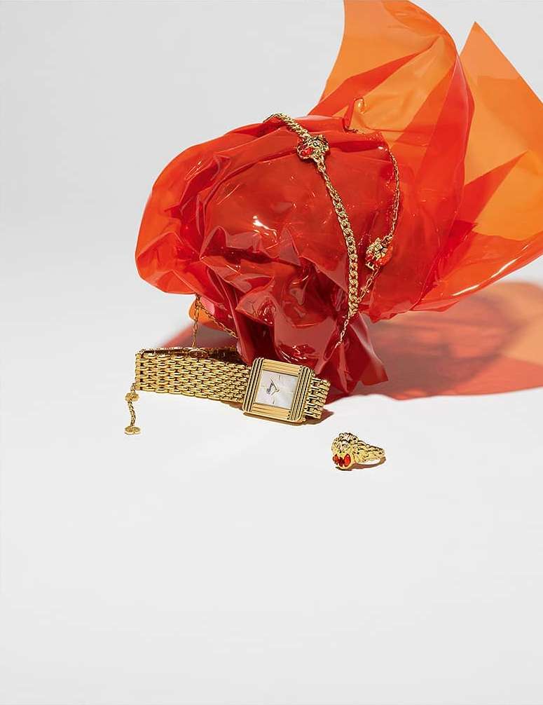 Still life shot of a gold watch and ring beside a gold chain on a white surface next to an orange plastic wrap.