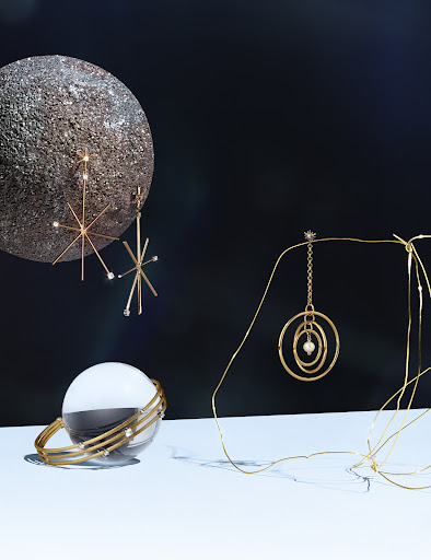 Still life shot of gold jewelry, including starburst earrings, a bangle bracelet, and a circular pendant, against a dark background.