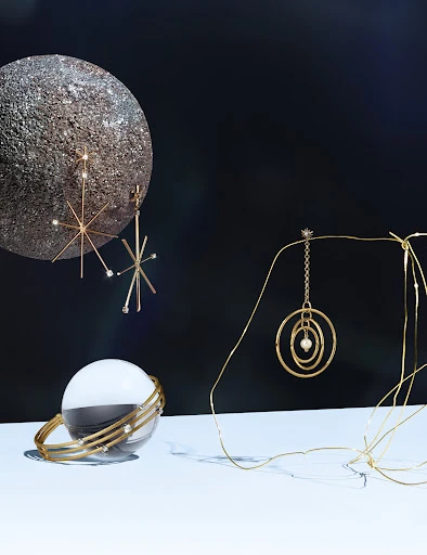 Still life shot of gold jewelry, including starburst earrings, a bangle bracelet, and a circular pendant, against a dark background.