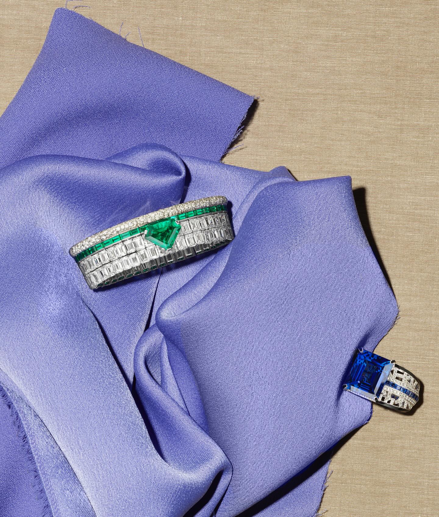 Close-up of two rings, one with an emerald and one with a sapphire, resting on soft, light purple fabric with a neutral background.