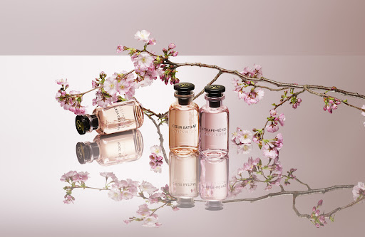 Three pink perfume bottles are arranged with cherry blossom branches on a reflective surface with a pink and white background.