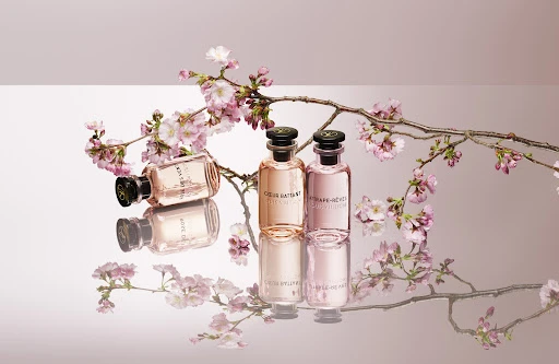 Three pink perfume bottles are arranged with cherry blossom branches on a reflective surface with a pink and white background.