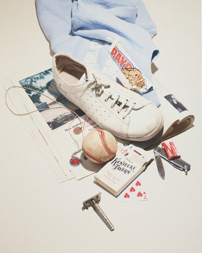 Flat lay arrangement of white sneakers, baseball, playing cards, razor, and miscellaneous personal items on an off-white background.