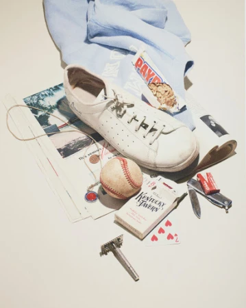 Flat lay arrangement of white sneakers, baseball, playing cards, razor, and miscellaneous personal items on an off-white background.