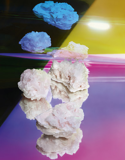 Geological formation of minerals in pink, blue, and yellow hues on a reflective surface, illuminated by vibrant light.