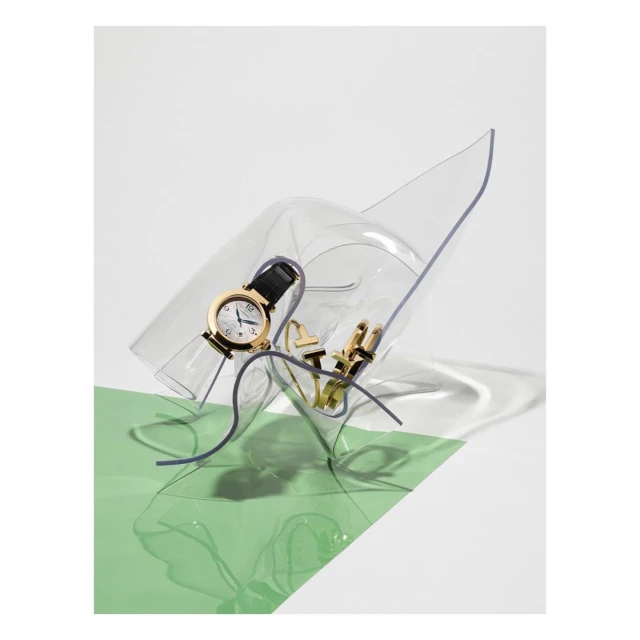 Still life shot of a gold watch with a black band on top of a clear, sculptural piece, with gold paper clips and a green backdrop.