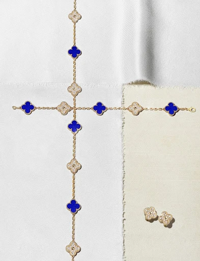 Gold necklace and earrings featuring four-leaf clover motifs in blue and white set against a backdrop of textured neutral fabric.