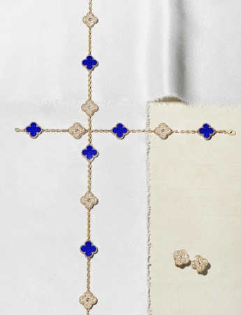 Gold necklace and earrings featuring four-leaf clover motifs in blue and white set against a backdrop of textured neutral fabric.
