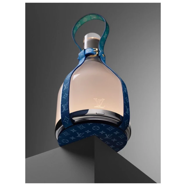 Elevated view of a Louis Vuitton lantern-style object featuring blue patterned leather straps and a glowing center, presented on a gray surface.