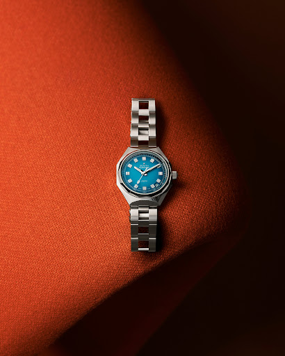 Wristwatch with a silver band and a teal face displayed on a textured orange background. The watch is positioned centrally in the frame.