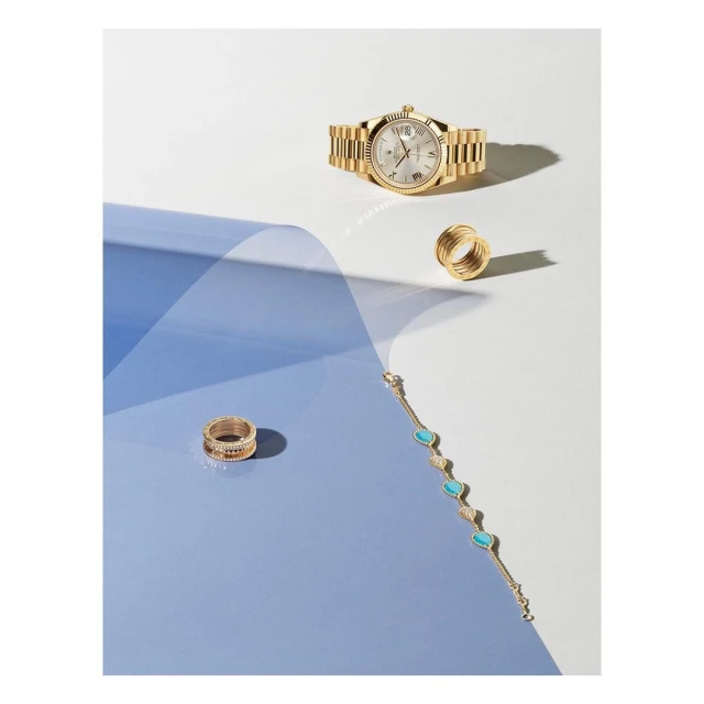Elevated studio shot of a gold wristwatch, gold rings, and gold bracelet with turquoise stones, all placed on a surface split blue and white.