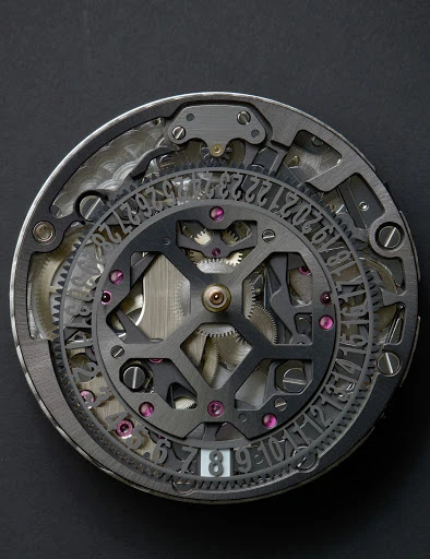 Close-up of an intricate watch mechanism featuring visible gears, screws, and number engravings under studio lighting against a dark background.