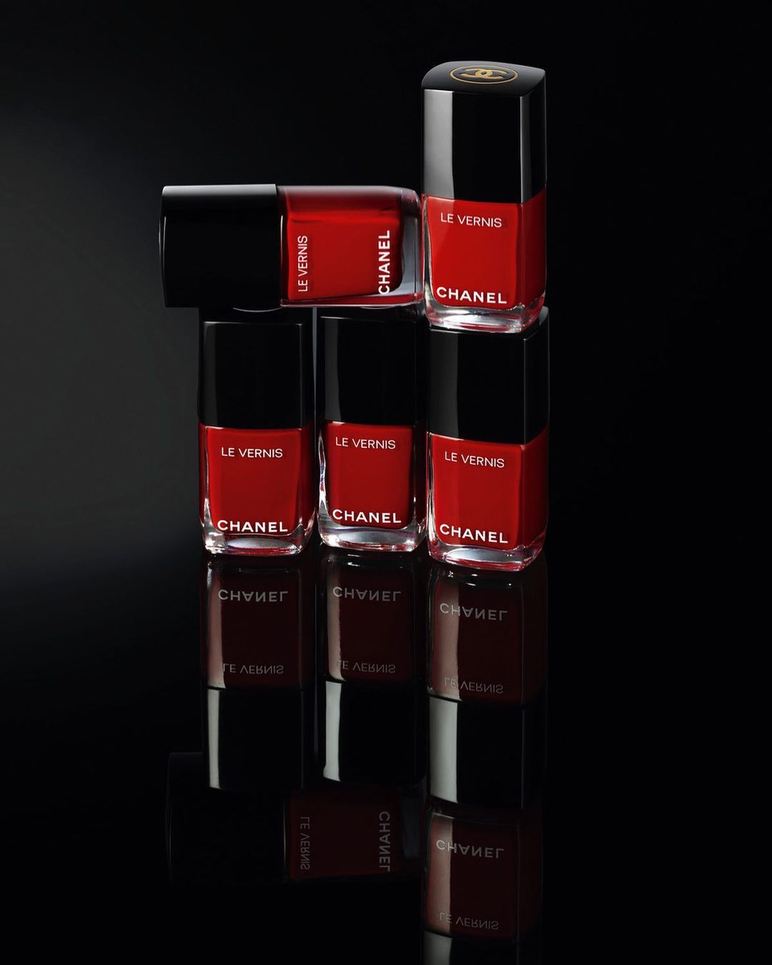 Stack of six red Chanel nail polish bottles with black caps, arranged on a reflective black surface in a beauty still life.