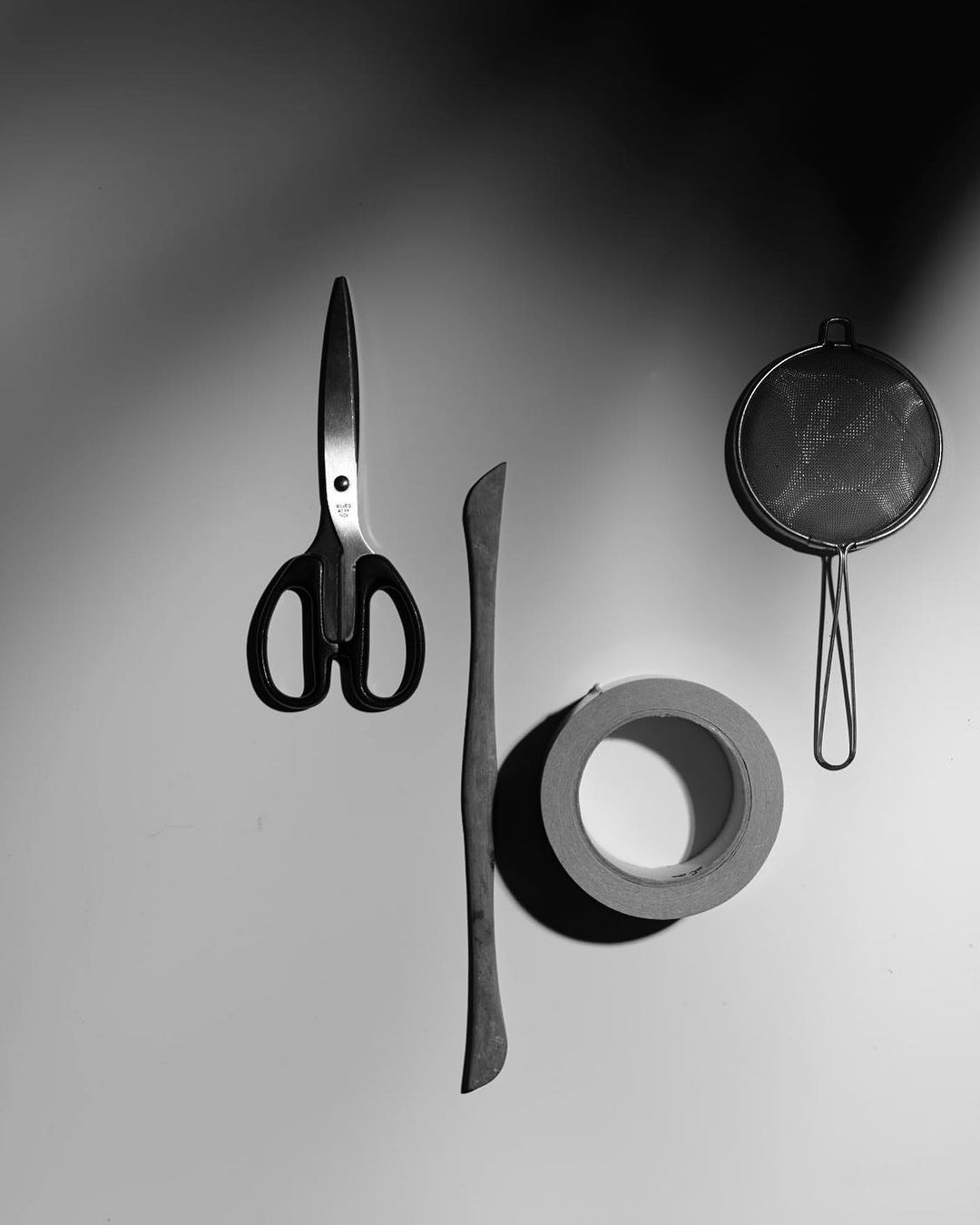 Monochromatic still life featuring scissors, wooden tool, tape roll, and strainer against a gradient background with soft lighting.