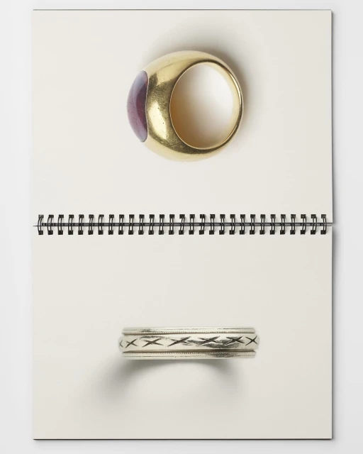 Two rings, one gold with a purple stone and one silver with a decorative band, sit on a white spiral-bound notebook.