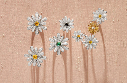 Seven flower-shaped rings with diamond petals and colorful gem centers are arranged on a textured, light pink surface.