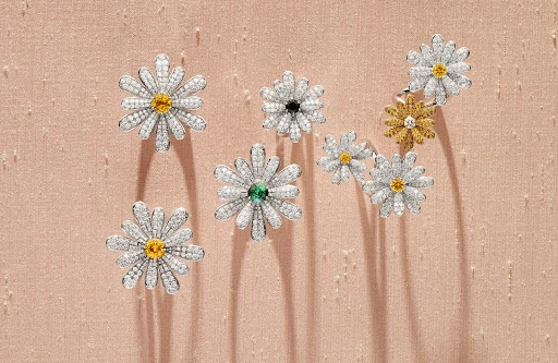 Seven flower-shaped rings with diamond petals and colorful gem centers are arranged on a textured, light pink surface.