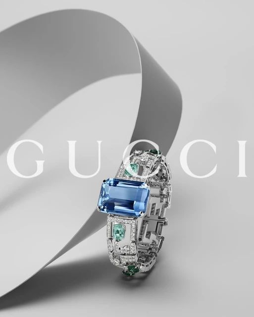 Gucci jewellery advertisement featuring a cuff-style bracelet with a large rectangular blue gemstone and smaller green gemstones on a gray background.