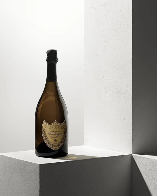 Dom Perignon champagne bottle on white concrete ledge, set against a brightly lit white background with geometric shadow pattern.