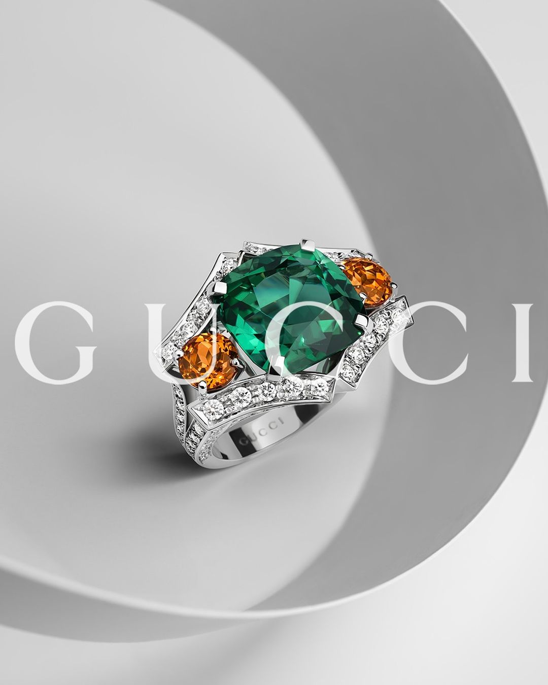 Close-up of an ornate silver ring featuring a large green gemstone flanked by smaller orange stones, set against a soft gray background.