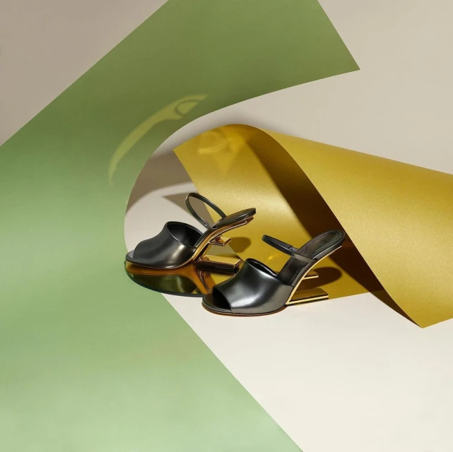 Two black, strappy, high-heel sandals with gold accents, displayed on a surface with light green, white, and gold paper accents.